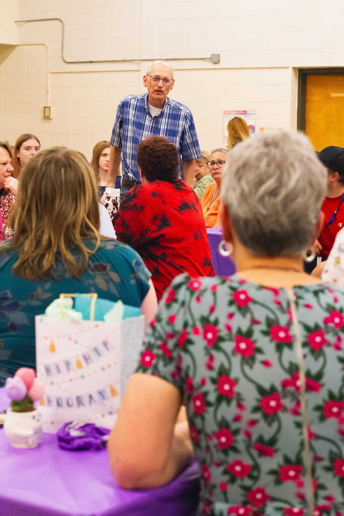 A longtime staff member stands and shares remarks with colleagues gathered around decorated tables during a retirement celebration in the Castleton Elementary School cafeteria.
