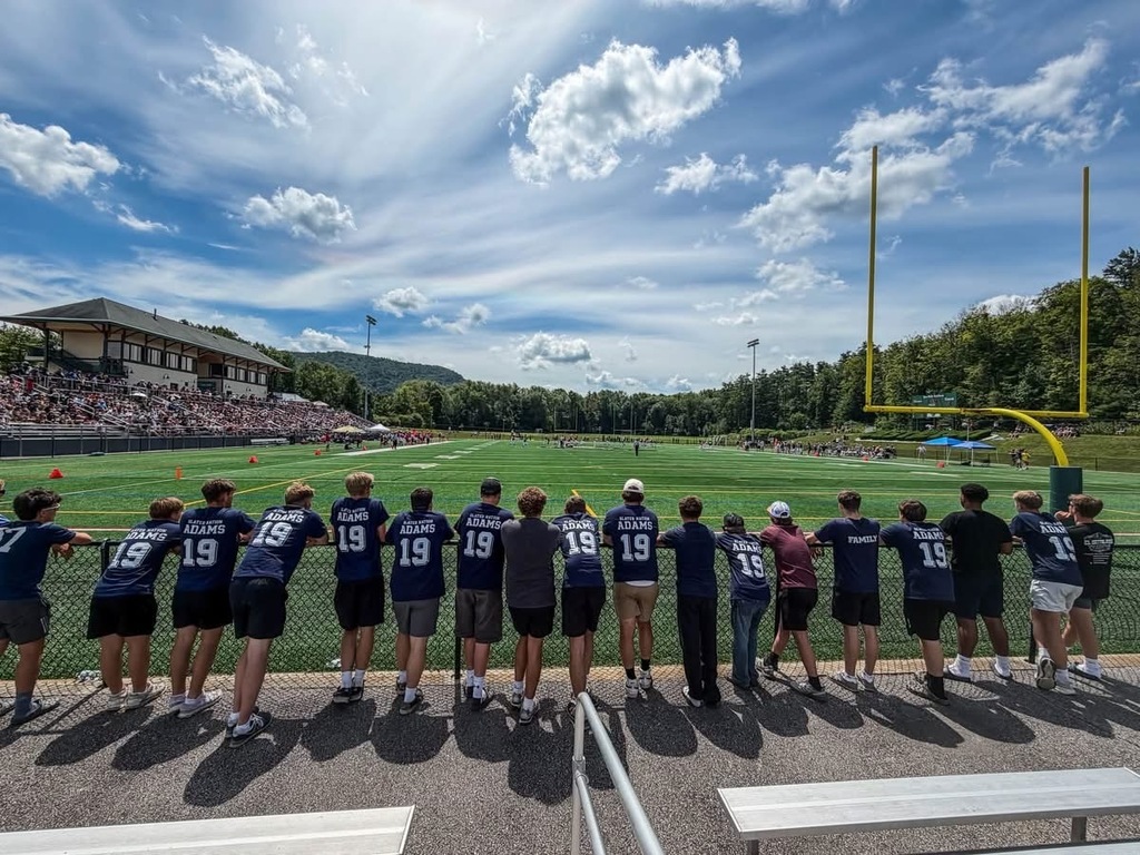 A wide view of the Maple Sugar Bowl football stadium shows spectators wearing matching "Adams 19" shirts lined up along a fence watching the game. The football field, goalposts, grandstands, and a bright blue sky with scattered clouds fill the background.