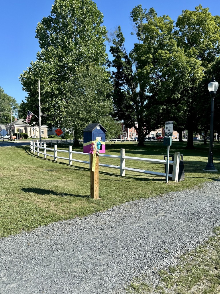 Wide view of the Slate Valley Community Mini Art Gallery installed near the north entrance of Fair Haven Park. The colorful gallery stands beside a white fence along a gravel path, surrounded by green grass, mature trees, and the village in the background under a bright blue sky.