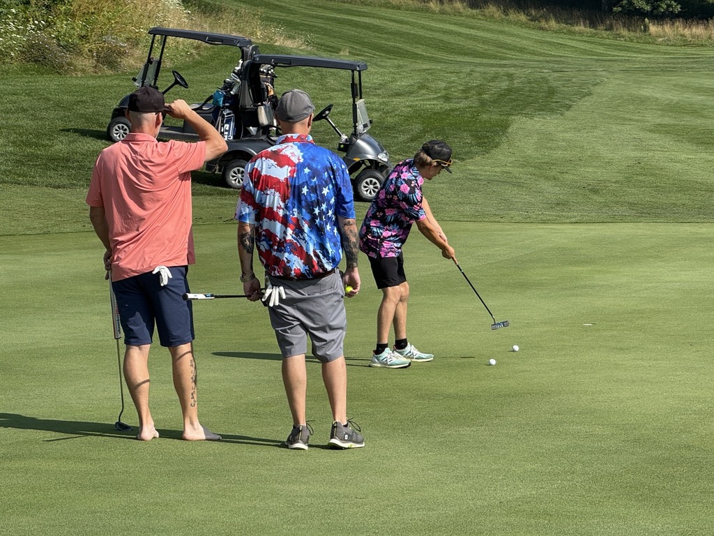 Three golfers stand on a putting green as one prepares to putt, with two golf carts and the course fairway visible in the background.