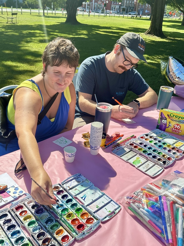 Two community members create miniature artwork at a table covered with watercolor paints, pencils, markers, and other art supplies.