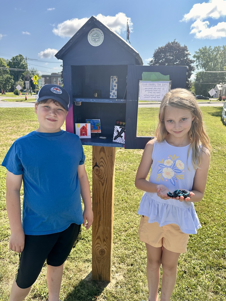Two children stand beside the open Slate Valley Community Mini Art Gallery, which displays several small pieces of artwork.