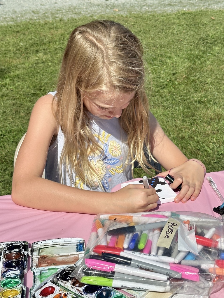 A young girl concentrates on creating a miniature drawing at an outdoor art table filled with paints and markers.