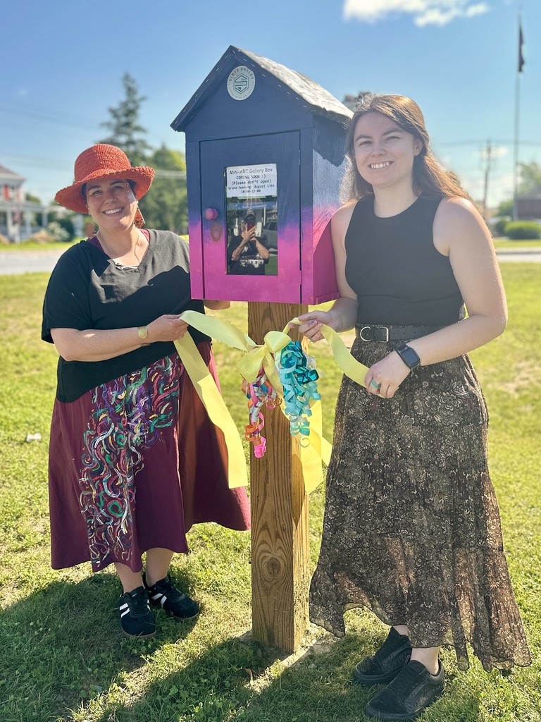 Two Slate Valley art teachers smile beside the Mini Art Gallery while holding the colorful ribbon from the opening ceremony.