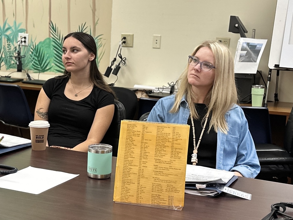 Two new Slate Valley educators sit at a conference table during New Teacher Orientation, listening to a presentation with orientation materials and beverages in front of them.