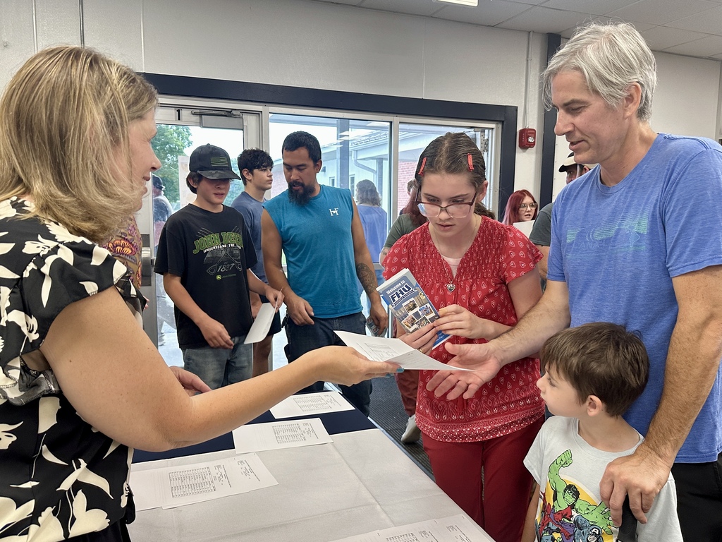 A staff member welcomes a student and family and hands the student a schedule at an Open House check-in table.
