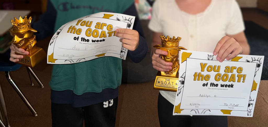 Two students hold gold G.O.A.T. (Greatest of All Time) trophies and certificates recognizing them as “G.O.A.T. of the Week.” The certificates are dated August 28, 2026.