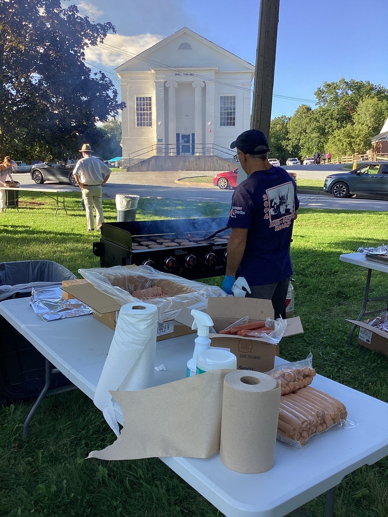 A person cooks burgers on an outdoor grill during a community gathering, with food-preparation tables in the foreground and the Orwell Town Hall visible across the street.