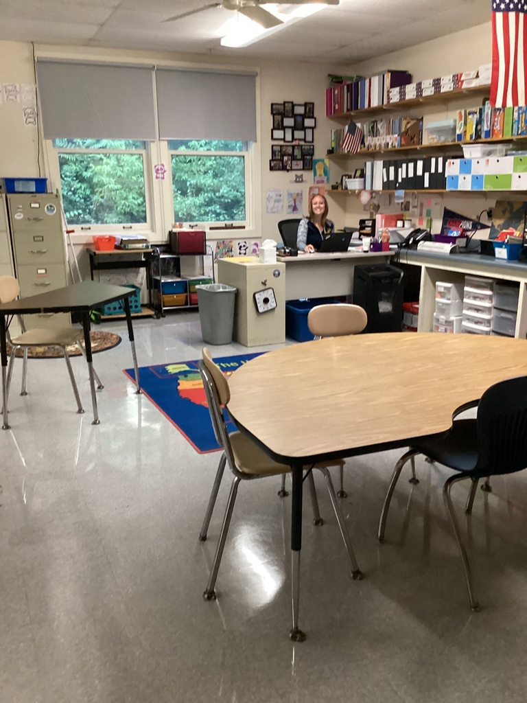 A staff member smiles from behind a desk in a bright, organized classroom featuring student tables, shelves of books and supplies, and American flags.