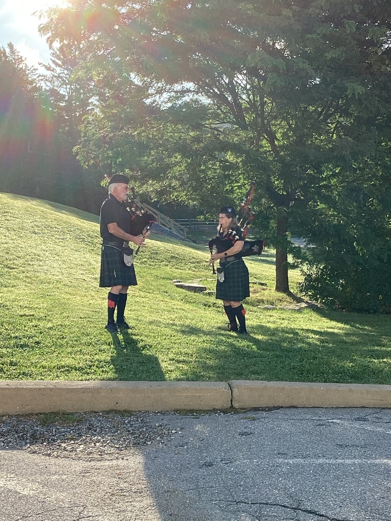 Two bagpipers in traditional kilts perform outdoors on a grassy area in the morning sunlight.