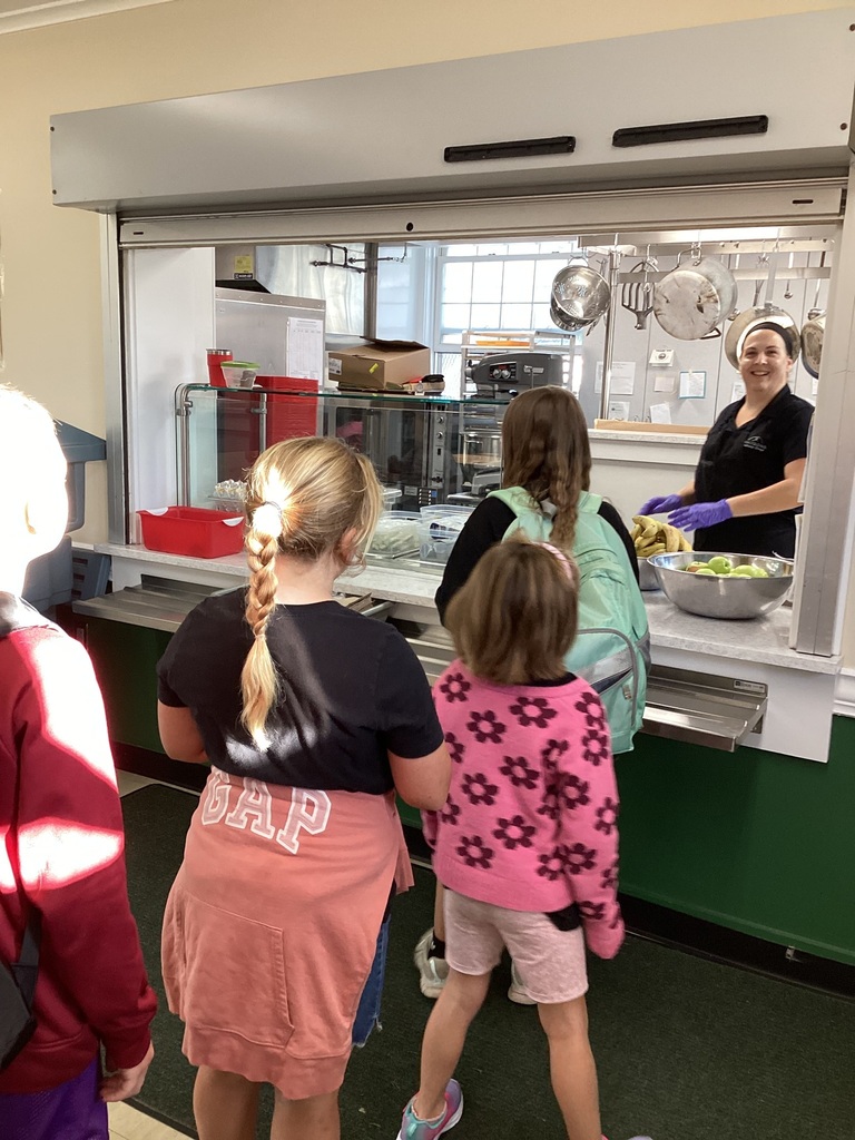 Students line up at the school cafeteria serving window as a smiling food service staff member prepares to serve fresh fruit.