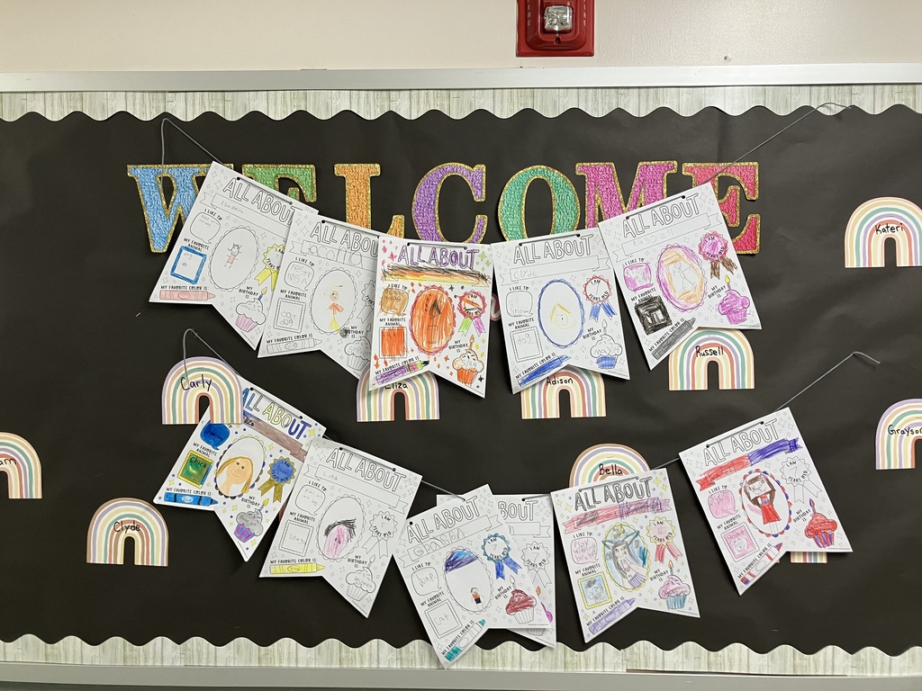 A classroom bulletin board titled "WELCOME" featuring strings of student-decorated "ALL ABOUT ME" pennants and pastel rainbow name tags.