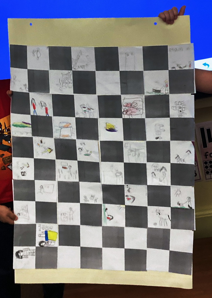 A handmade classroom quilt created by Mrs. McCrea’s first grade students. The large paper quilt features alternating gray and white squares, with students’ colorful drawings filling many of the white squares. Two people hold up the finished project for display.