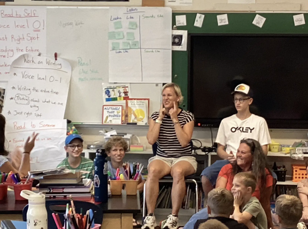 Students and adults smile and interact during a classroom group activity. An adult seated at the front gestures while talking, as students gathered around her listen and respond.