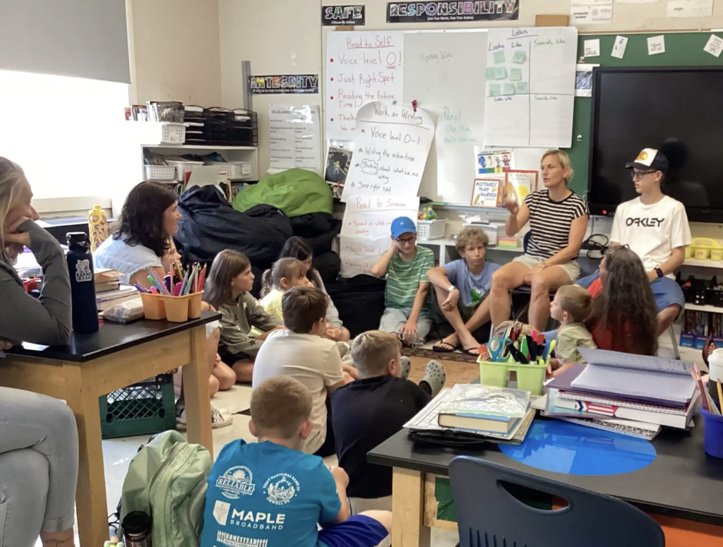 Students and adults gather in a classroom for a group discussion, with students seated on the floor and adults seated among them. Classroom posters, books, supplies, and a whiteboard are visible in the background.