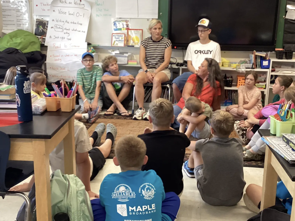 Students sit together on the classroom floor while several adults join them for a group conversation. Participants are gathered in a circle near the front of the classroom.