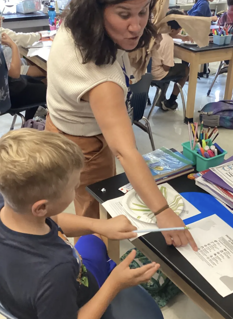 Educator works one-on-one with a student at a classroom desk, pointing to the science worksheet as they discuss the toothpaste investigation.