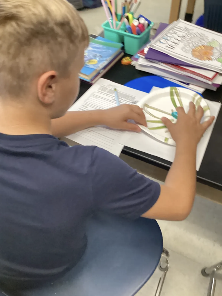 Student seated at a classroom desk examining a small sample of blue toothpaste on a paper plate as part of a science investigation.