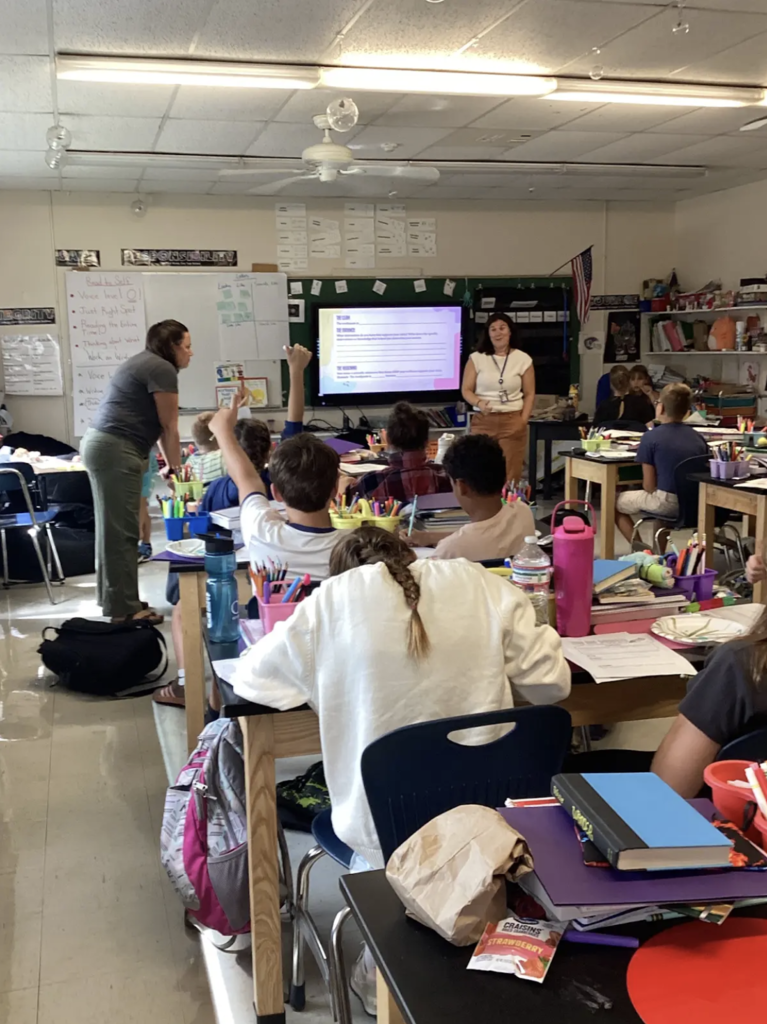 Students participate in a classroom science lesson while two educators lead a discussion at the front of the room. One student raises a hand to participate.