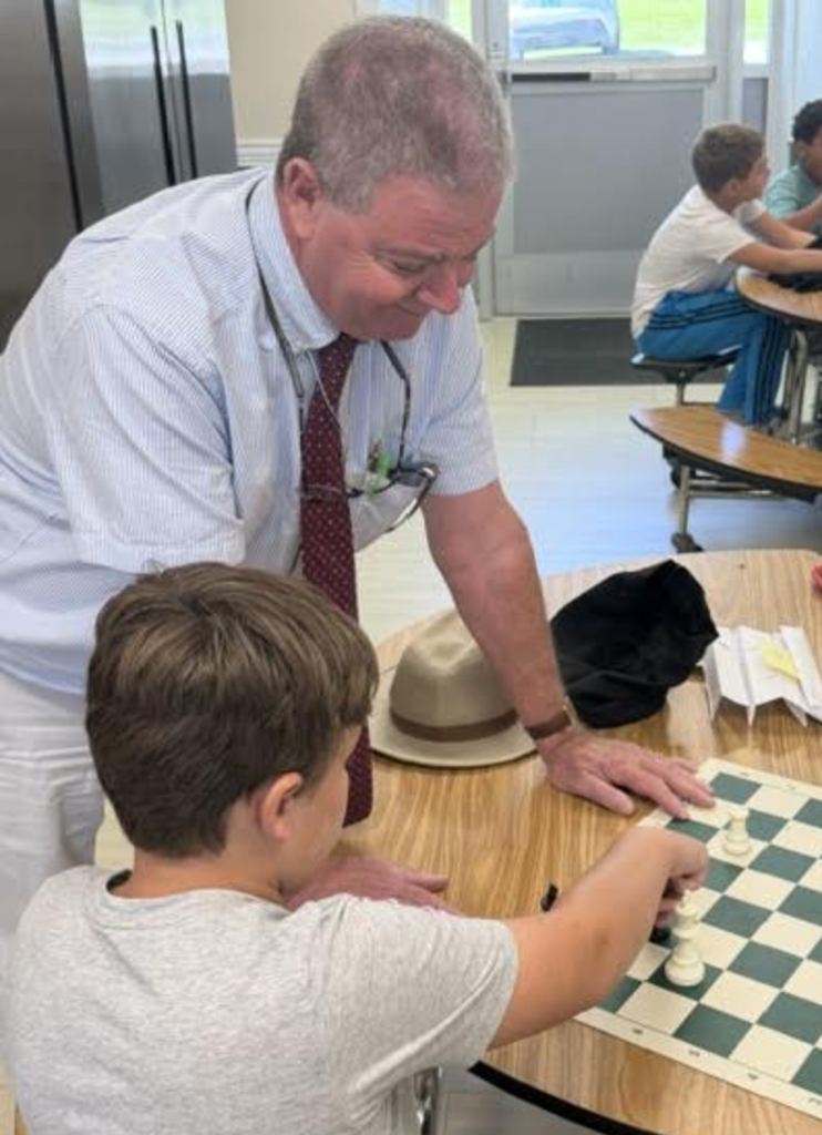 An adult leans over a cafeteria table and watches as a student moves a piece on a chessboard. Other students sit at tables in the background.