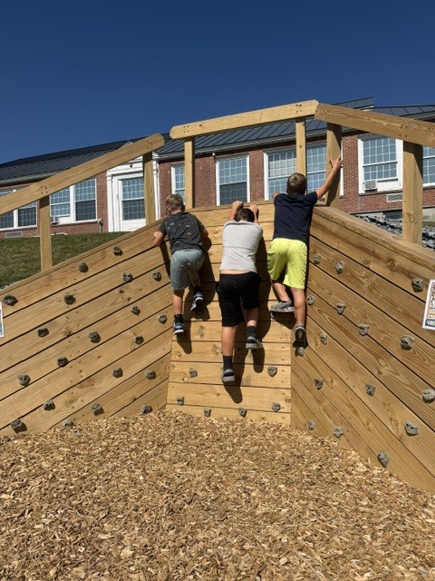 Three students climb a large wooden rock-climbing structure on a school playground on a sunny day.
