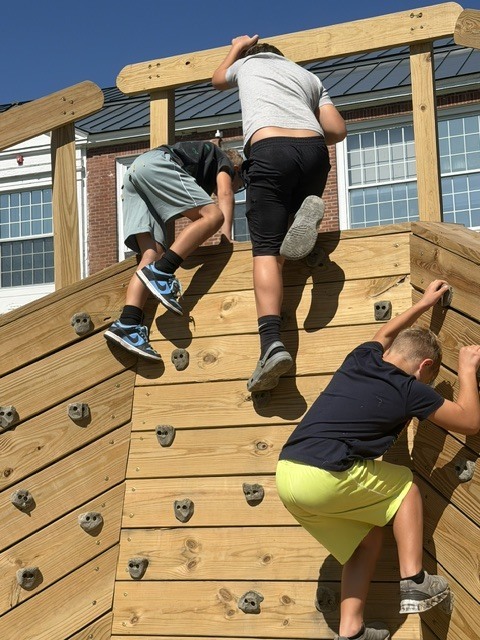 Close-up of three students using handholds and footholds to climb different sections of a wooden playground climbing wall.