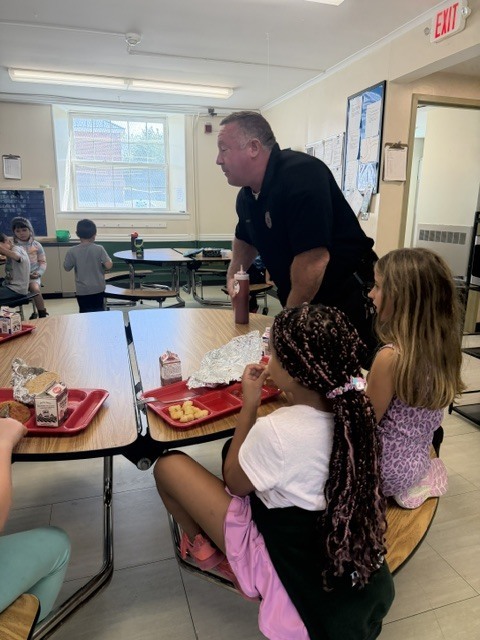 A law enforcement officer talks with a group of elementary students seated together at a cafeteria table during lunch.