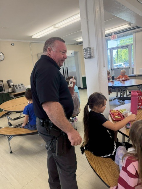 A uniformed law enforcement officer visits with elementary students as they eat lunch in the school cafeteria.