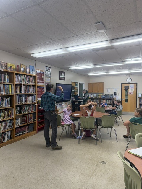A teacher stands in a school library presenting a lesson on a display screen to students seated at tables.