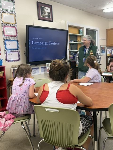 Elementary students sit at tables in a school library, working on activities while a presentation titled "Campaign Posters" displays on a TV screen.