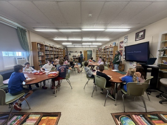 Students sat at a table in a library look toward a monitor displaying a presentation on campaign posters, while a teacher assists nearby.