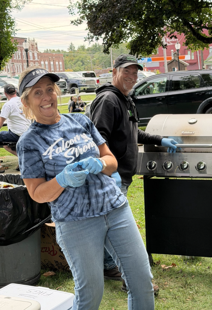 A smiling woman in a tie-dye shirt and visor wearing gloves stands near a outdoor grill operated by a man wearing a black hoodie and cap.