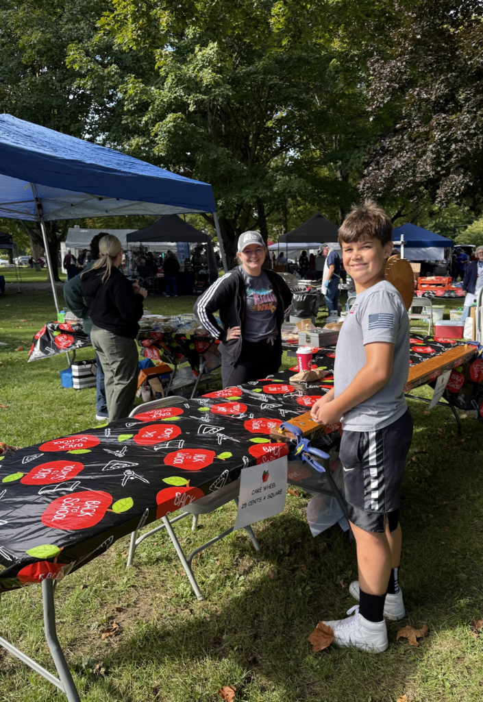 A young boy and a woman stand behind a long table set up for a cake wheel game at the park.