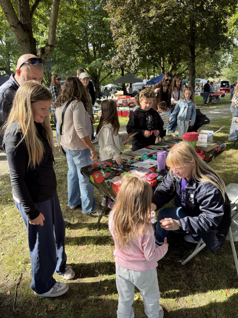 A woman kneels to help a young child at an activity table while other kids play outdoor carnival games nearby.