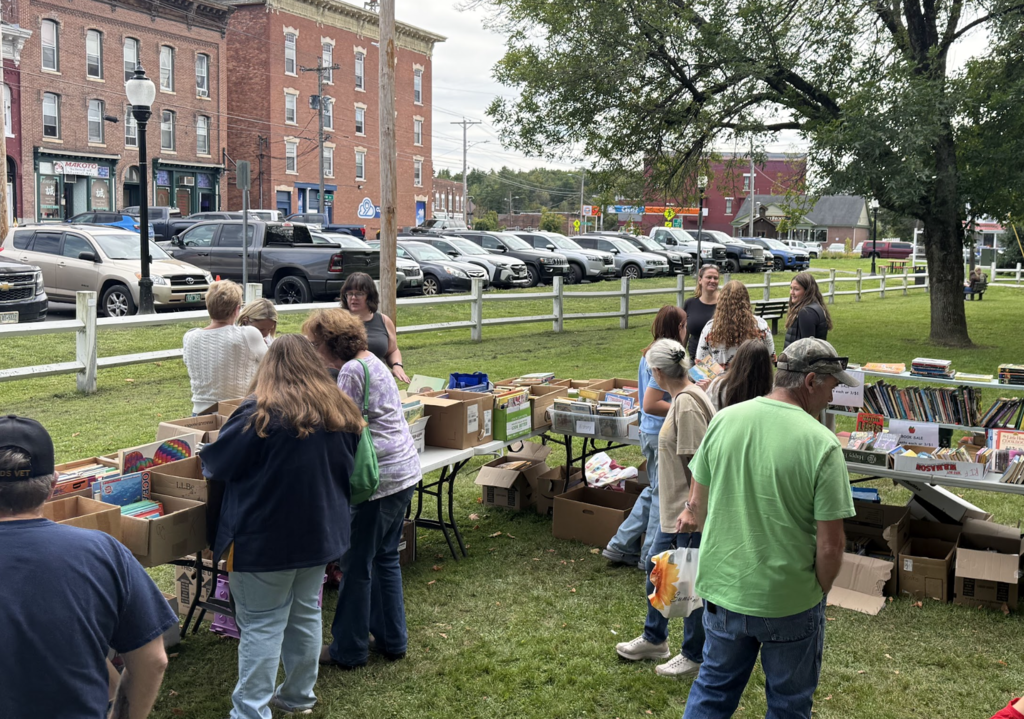 Community members browse through boxes and displays of books at an outdoor book sale table in the park.