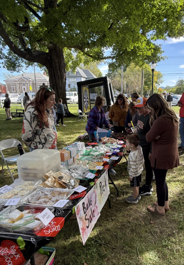 Community members and children gather around a table filled with baked goods for a Pre-K bake sale.
