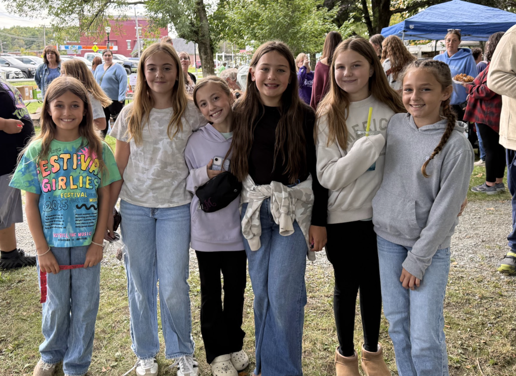 Six smiling young girls stand together outdoors posing for a photo at Applefest.