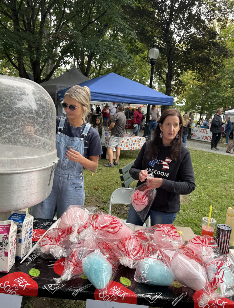 A woman in denim overalls spins cotton candy while another volunteer bags pink cotton candy at a festival booth.