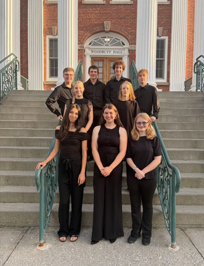 A group photo of nine student musicians, dressed in black performance attire, standing on the steps of Woodruff Hall. They are smiling at the camera. Woodruff Hall, a brick building with white columns and large wooden doors, is in the background.