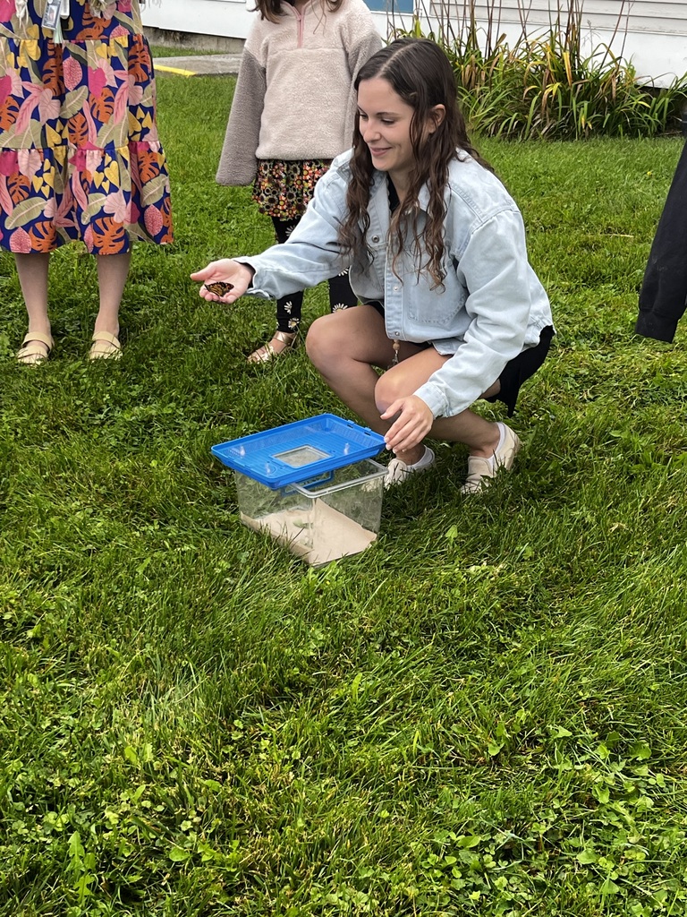 A young woman crouching in a grassy lawn, holding a butterfly on her outstretched hand next to an open clear container.