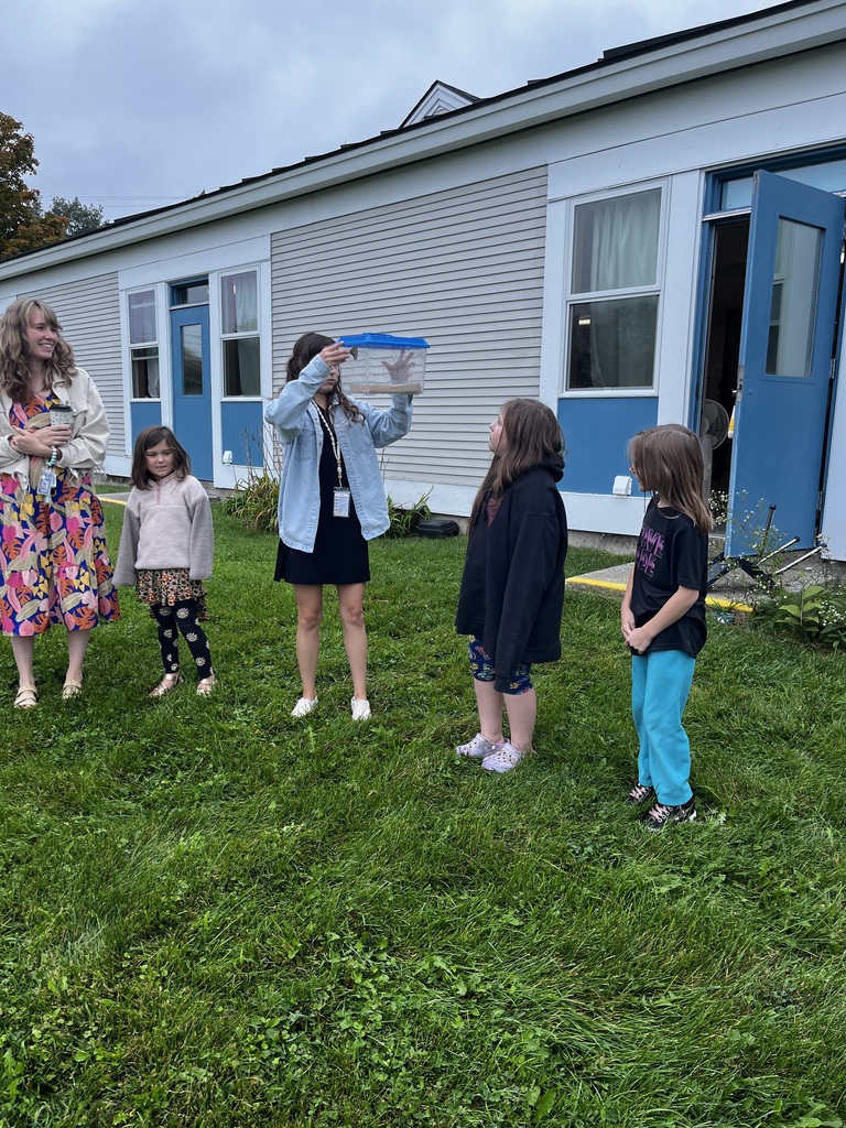 A teacher and children standing in a grassy yard outside a school, looking at a girl holding up a clear plastic creature container with a blue lid.