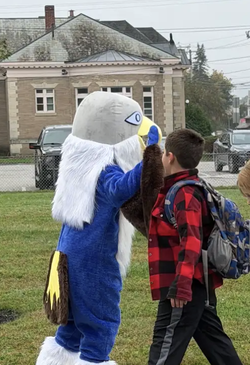 A student in a red and black plaid jacket gives a high-five to the falcon mascot outside on the school grounds.