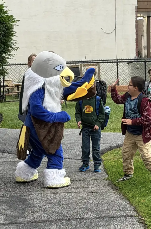 A group of elementary students wearing backpacks gather along a walkway outside to greet and high-five the falcon mascot.