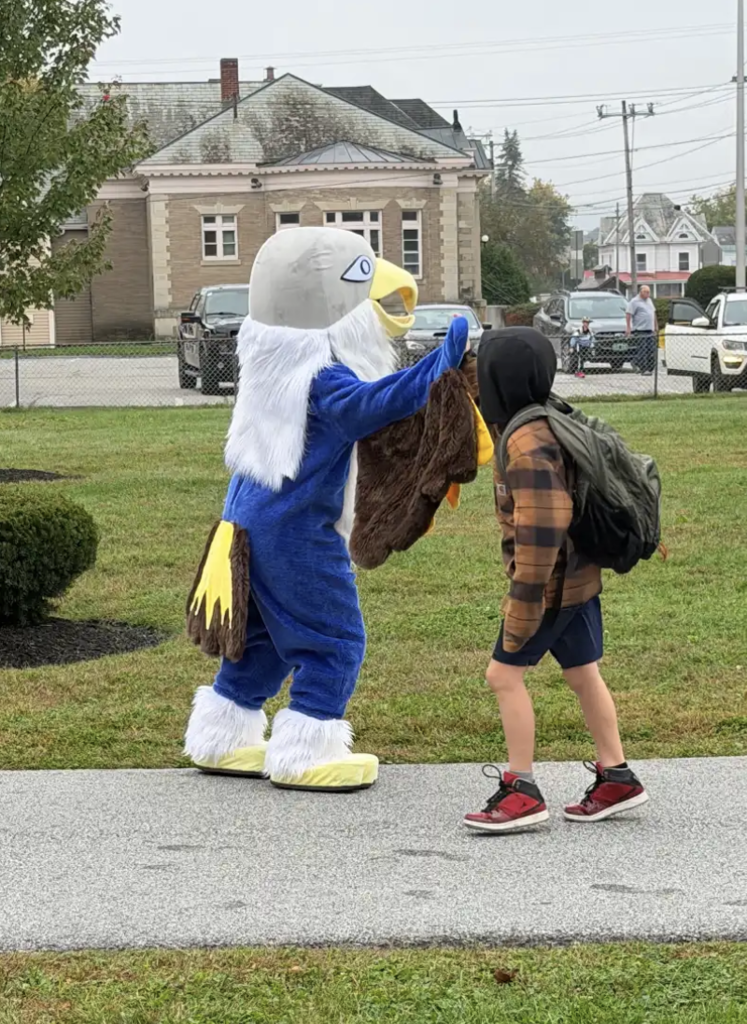The Fair Haven Grade School eagle mascot high-fives a young student wearing a jacket and backpack walking on a walkway outside the school building.