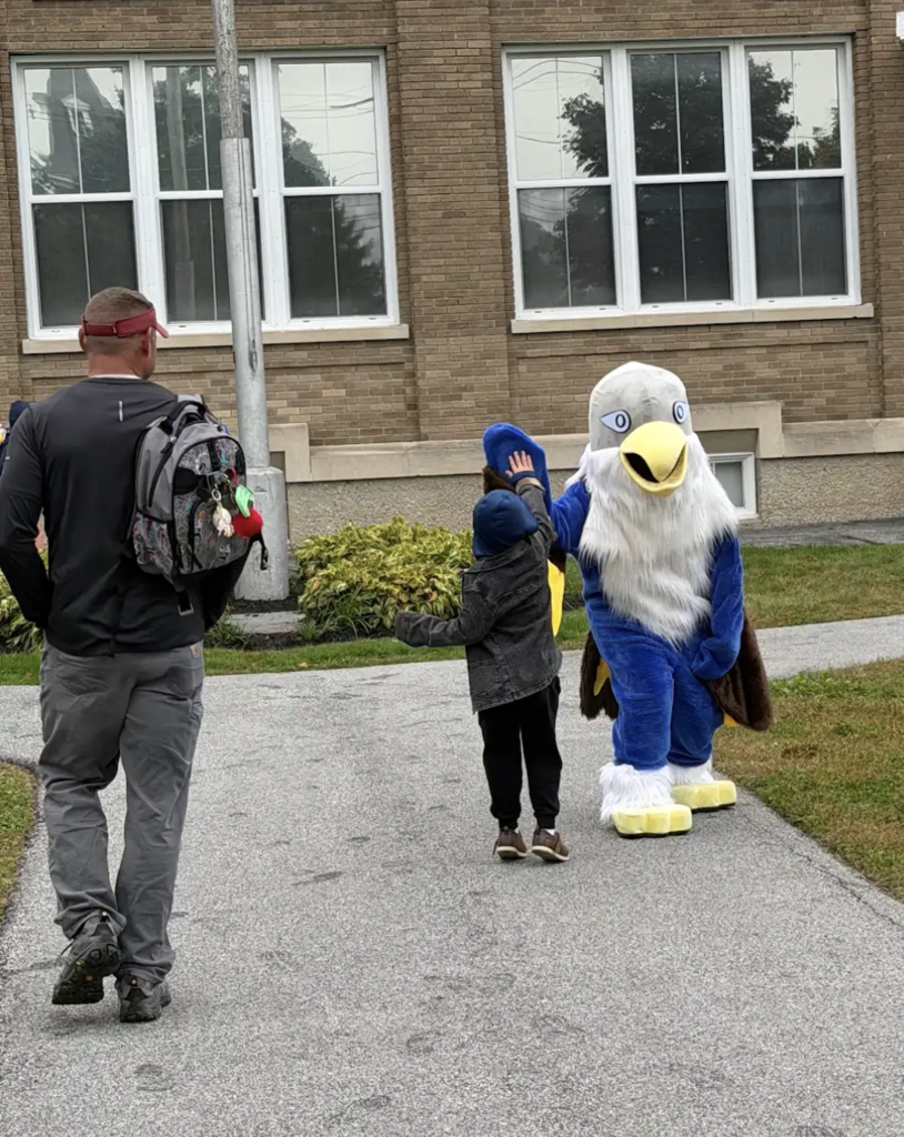 The Fair Haven Grade School mascot, a blue falcon with a white head and yellow beak, stands on a paved walkway in front of the school building with open wings welcoming students.