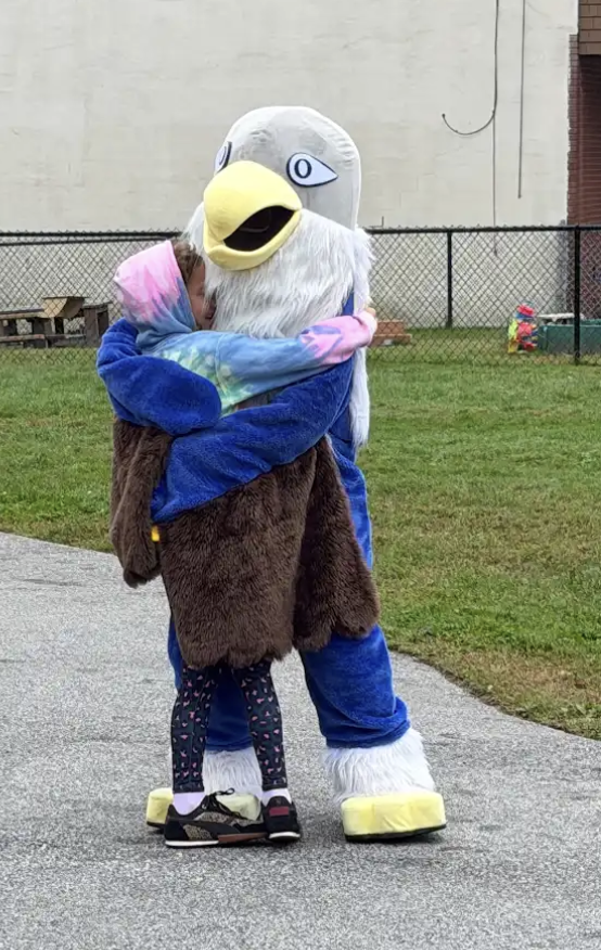 A young student wearing a colorful tie-dye hoodie gives a big hug to the school's falcon mascot.