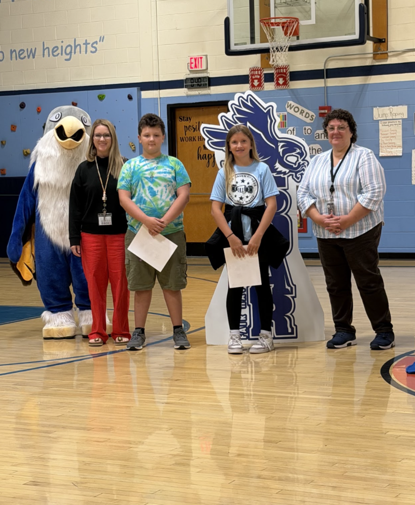 Two award-winning students stand on the gym floor flanked by two staff members and the falcon mascot.