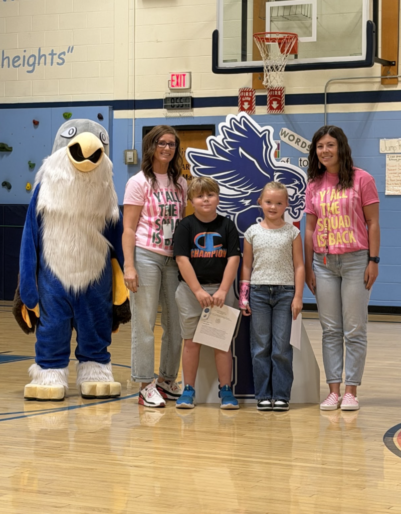 Two students holding award certificates pose on the gym court with two staff members in matching pink shirts and the falcon mascot.