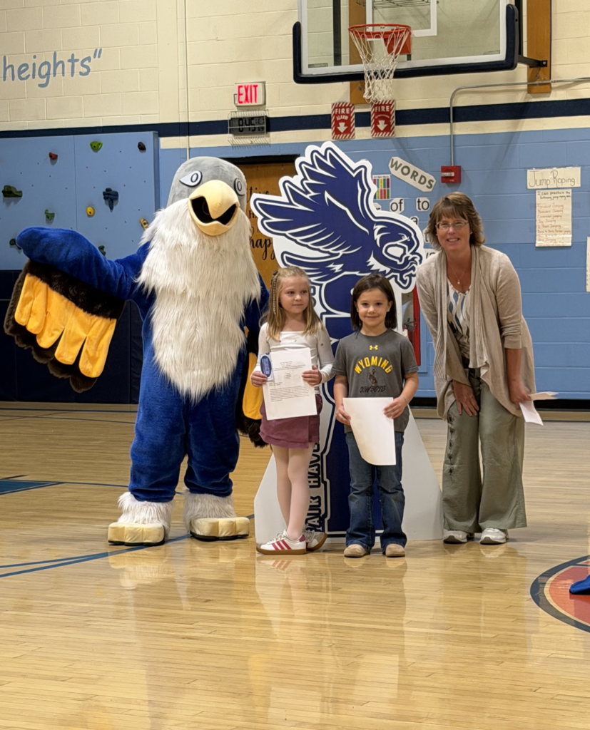 Two young students hold award certificates while standing with a staff member and the falcon mascot in the gym.