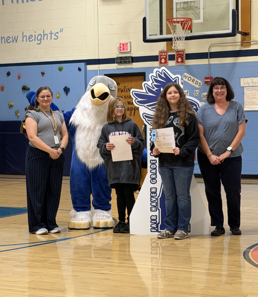 Two students display their award papers alongside two staff members and the falcon mascot on the gymnasium court.